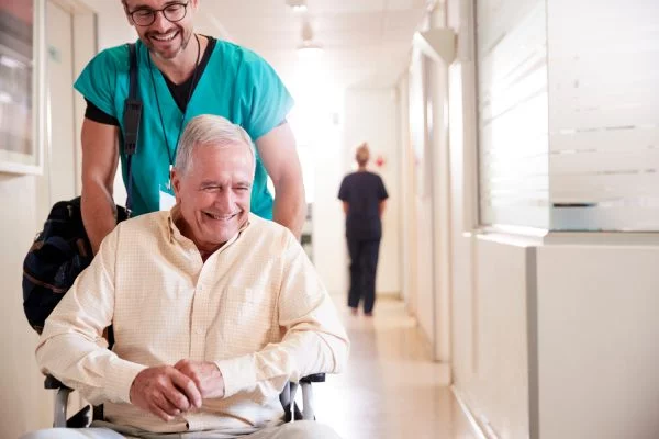 Male Orderly Pushing Senior Male Patient Being Discharged From Hospital In Wheelchair