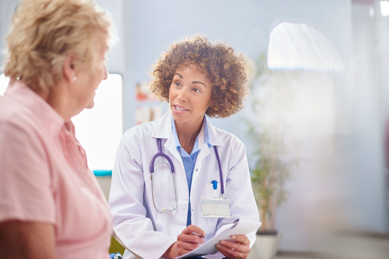 a female doctor sits and listens to a senior woman sitting down in a consultation room . They are sitting in a defocussed hospital clinic waiting room . The doctor is wearing a white coat and holding a prescription pad . She is smiling as they chat .