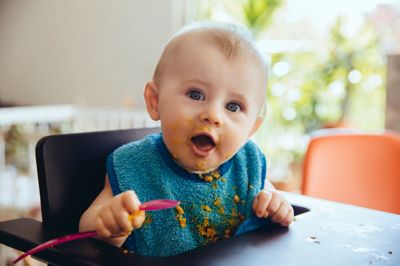 Baby boy with spoon and bib sitting in chair all happy after having had a meal